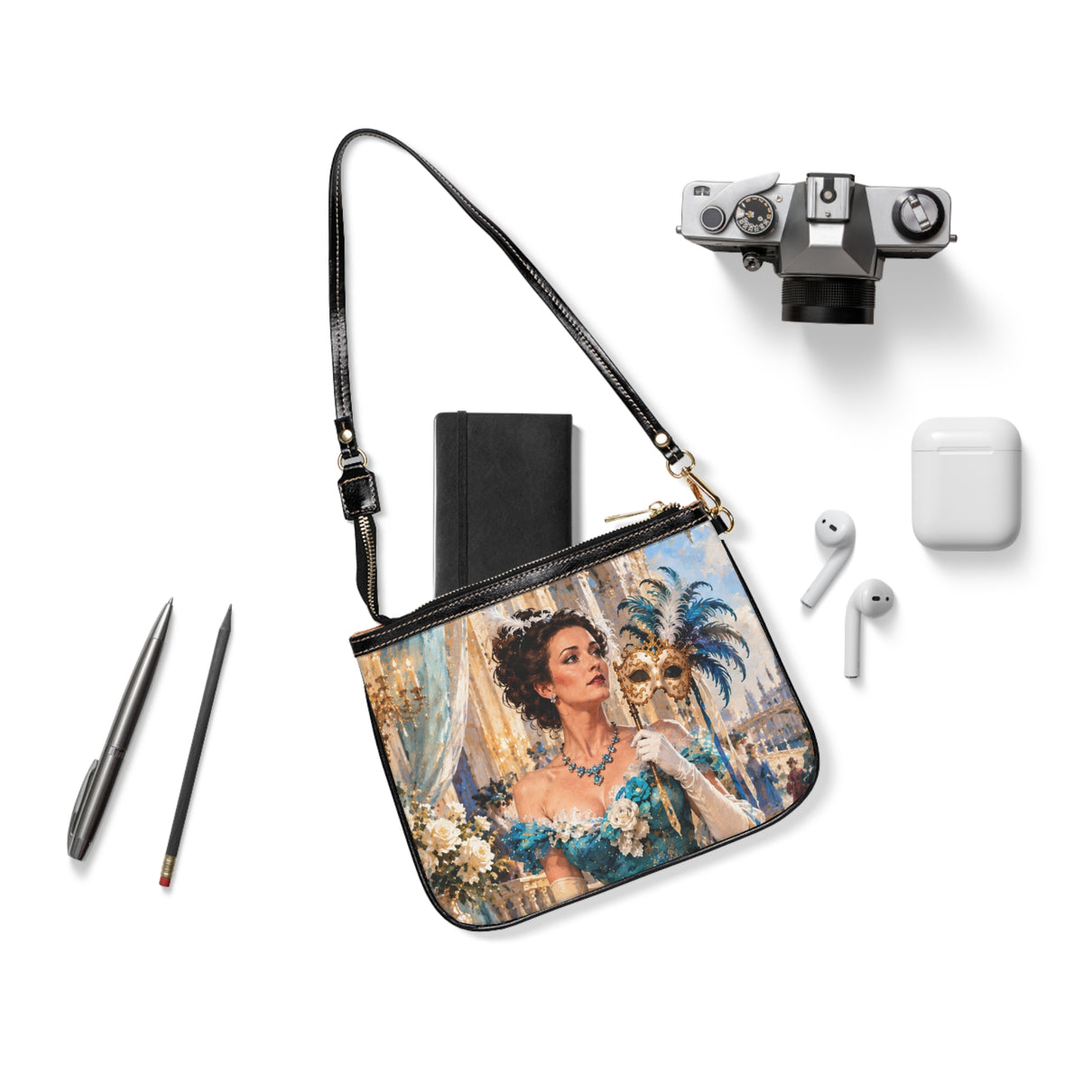 The Masquerade — A Venetian Dream small shoulder bag laid flat with its black strap beside a camera, notebook and earbuds.